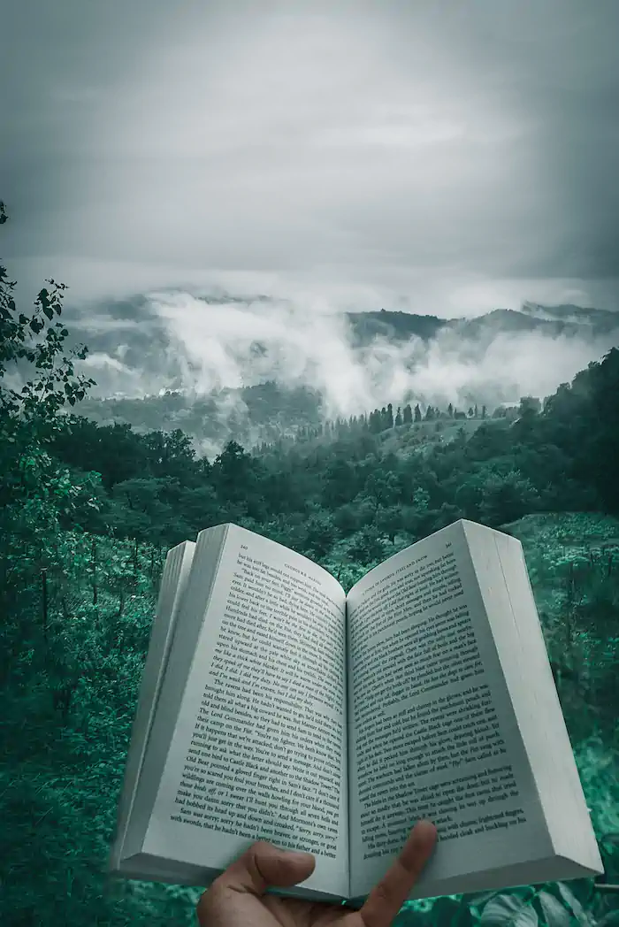 Image shows a hand holding a book open in the middle. In the background are mist covered low mountains.