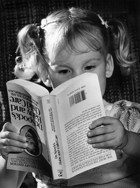 Black and white photo of a little girl reading Dr. Spock's Baby and Childcare. She is holding the book upside down.